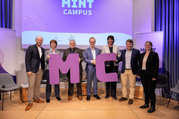 Sieben Personen stehen lächelnd auf einer Bühne und halten große violette Buchstaben mit dem Wort „MINT“ in den Händen. Auf einer Leinwand hinter ihnen wird das Wort „MINT-Campus“ angezeigt. Hintergrund und Dekoration sind überwiegend in Violett- und Weißtönen gehalten.