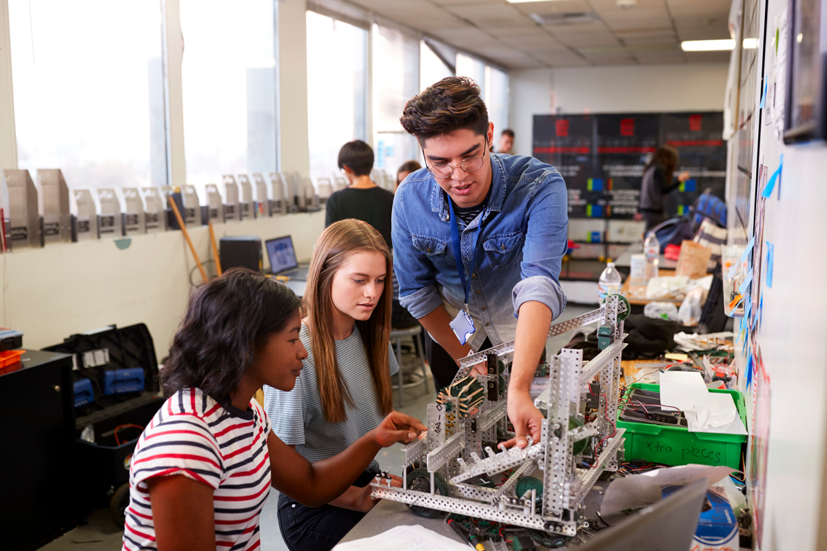 Drei Schüler in einem Klassenzimmer arbeiten an einem überfüllten Arbeitsplatz zusammen und konstruieren ein Robotergerät aus Metallteilen und Drähten. Zwei sitzende Schüler, einer in einem gestreiften Hemd und einer in einem grauen Hemd, beobachten, wie ein stehender Schüler in einem Jeanshemd einen Teil erklärt.