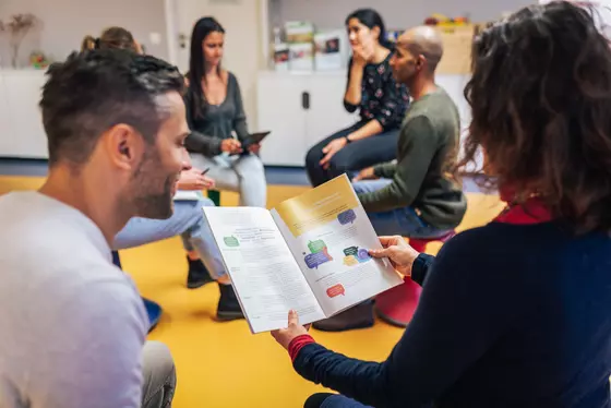 Eine Gruppe von Menschen sitzt im Kreis und diskutiert. Eine Person im Vordergrund hält eine Broschüre mit Diagrammen und Texten zu „Forschendes Lernen“ in der Hand und liest sie. Die Umgebung scheint eine gemütliche Innenumgebung mit sanfter Beleuchtung zu sein.