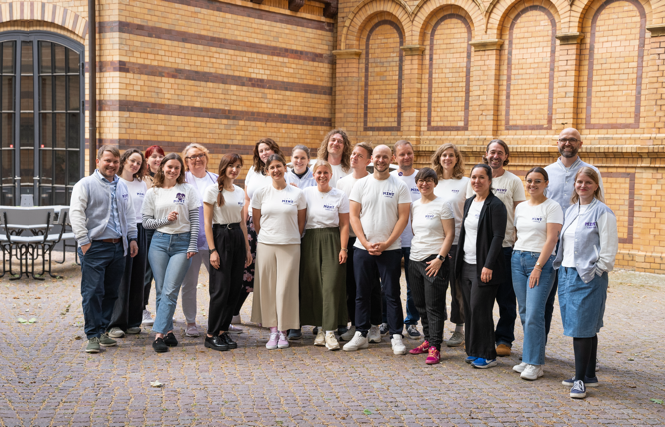 Eine Gruppe von 22 Personen steht gemeinsam vor einem Backsteingebäude auf dem MINT-Campus-Teamevent, alle sind leger gekleidet, einige tragen passende weiße T-Shirts mit Logo. Der Innenhof mit Kopfsteinpflaster ergänzt diesen Treffpunkt für digitale Weiterbildung.