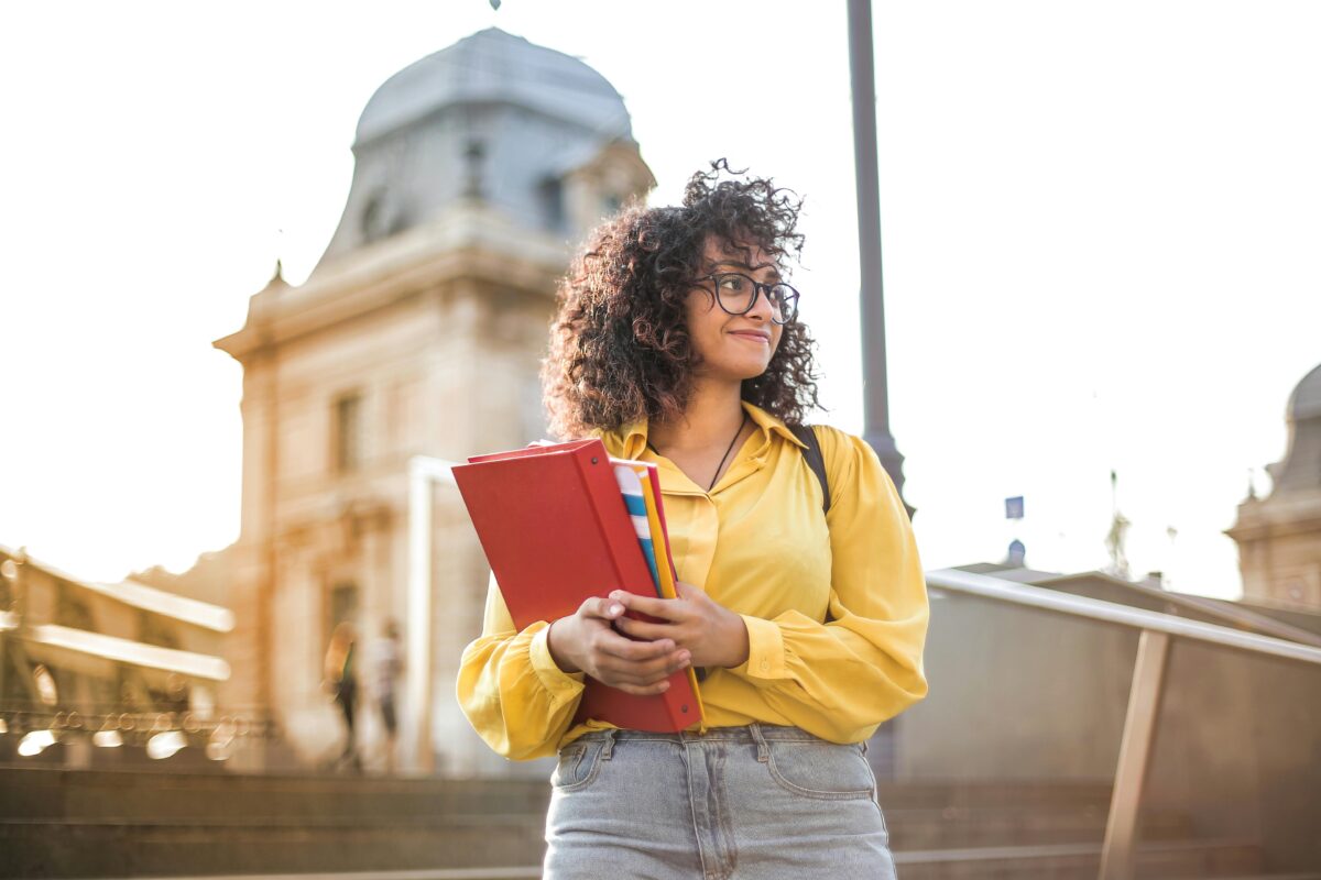 Ein junger Mensch mit lockigem Haar und Brille steht im Freien und hält einen roten Ordner und Bücher in der Hand. Er trägt ein gelbes Hemd und hell gewaschene Jeans. Im Hintergrund ist ein historisches Gebäude zu sehen und die Sonne scheint.