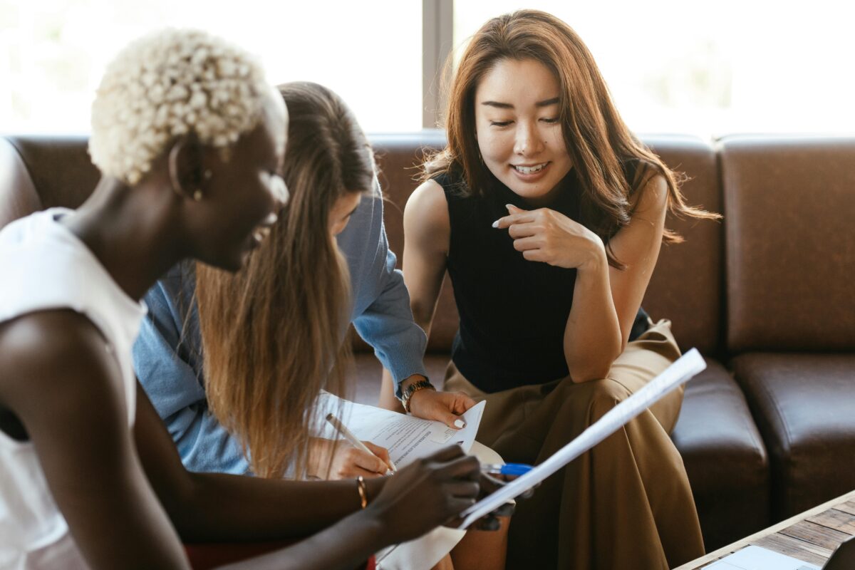 Drei Frauen sind in einem hellen Raum versammelt. Sie sitzen auf einem Sofa, betrachten Dokumente und diskutieren angeregt etwas. Eine Frau hält einen Stift in der Hand und scheint etwas auf dem Papier zu erklären oder darauf hinzuweisen.