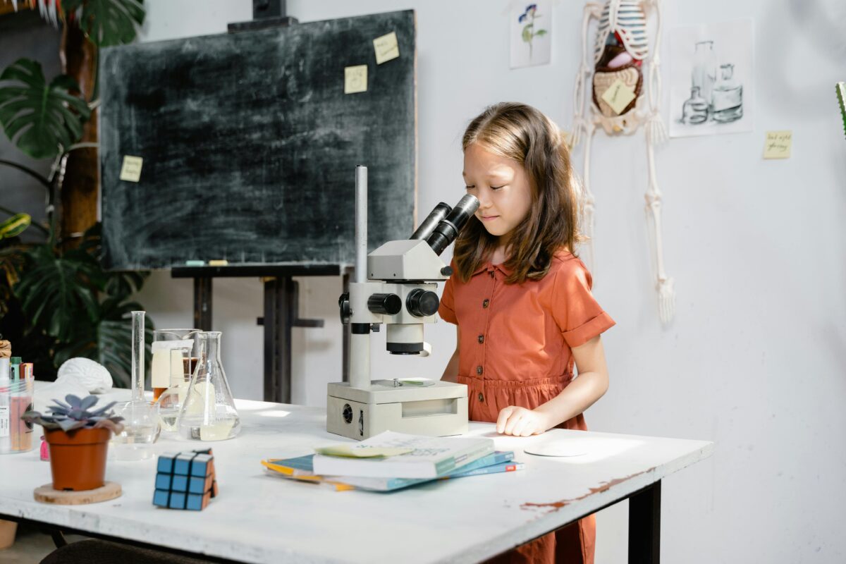 Ein junges Mädchen in einem orangefarbenen Kleid schaut durch ein Mikroskop auf einem mit Büchern, einem Zauberwürfel und Laborgeräten vollgestopften Tisch. Hinter ihr ist eine Tafel mit Haftnotizen und eine Wand mit verschiedenen wissenschaftlichen Diagrammen und Zeichnungen.