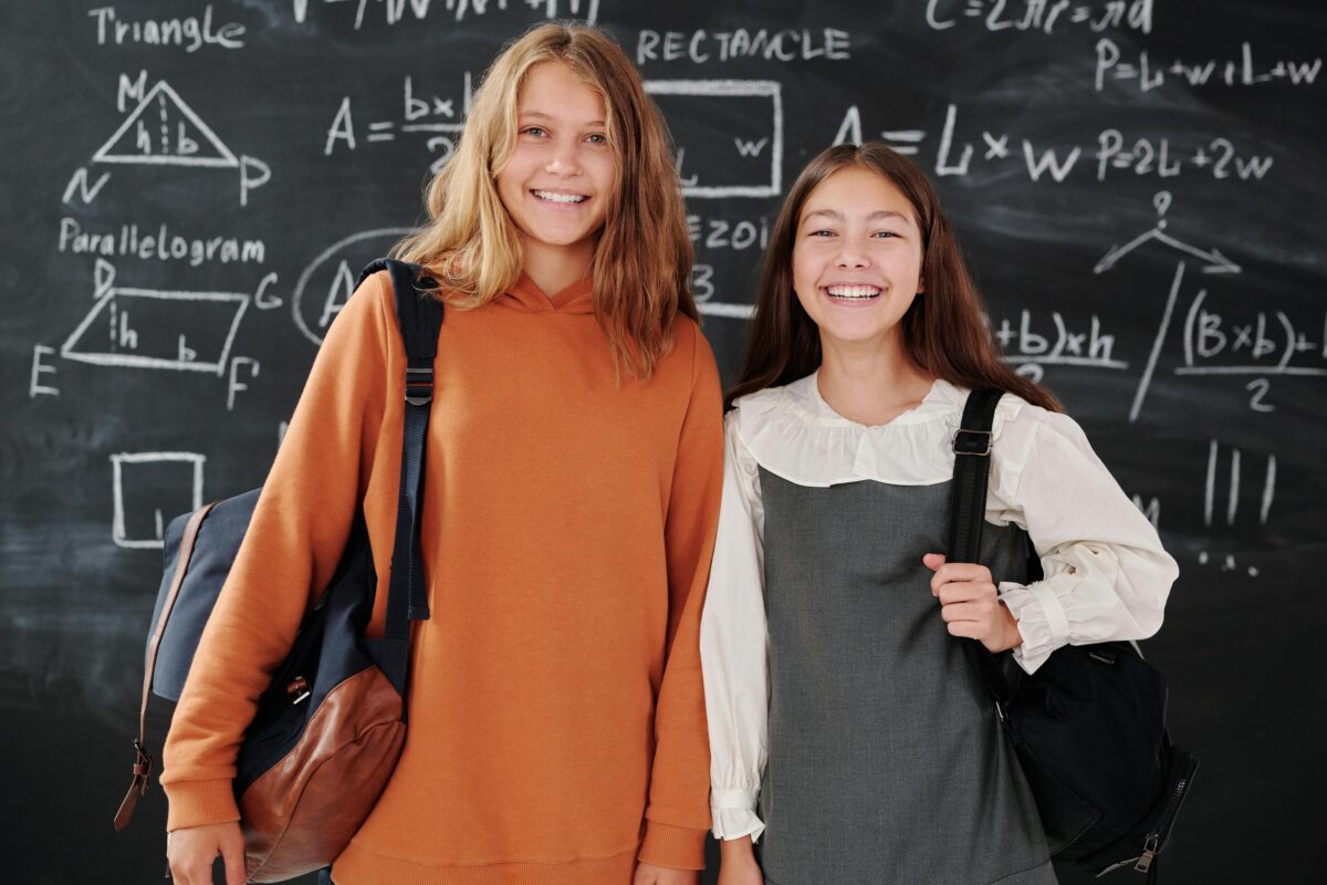 Zwei junge Schülerinnen stehen vor einer Tafel voller mathematischer Formeln und Diagramme. Beide lächeln, tragen Rucksäcke und sind leger gekleidet. Die Schülerin links trägt einen orangefarbenen Kapuzenpullover, während die Schülerin rechts eine weiße Bluse und ein graues Kleid trägt.