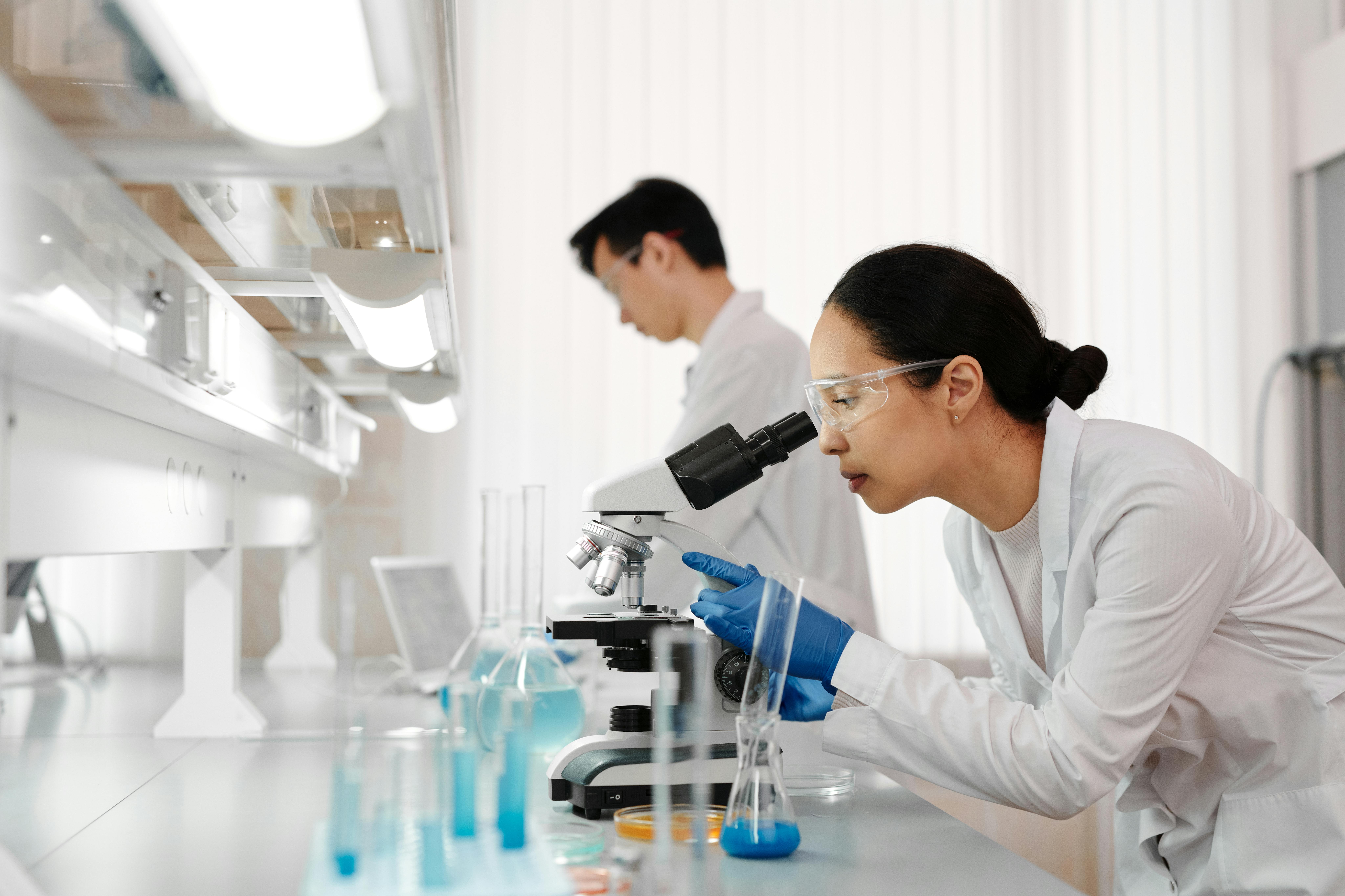 Eine Frau im Laborkittel und mit Schutzbrille schaut in einem Labor durch ein Mikroskop. Im Hintergrund arbeitet ein Mann im Laborkittel mit Laborgeräten. Der Arbeitsbereich ist hell und mit verschiedenen wissenschaftlichen Instrumenten und Glaswaren gefüllt.