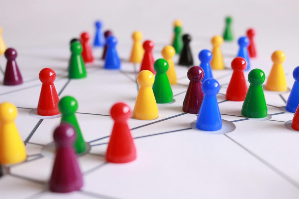 Bunte Plastikspielsteine, darunter rot, gelb, grün, blau, violett und schwarz, werden auf einem verbundenen Netzwerkdiagramm platziert. Das Diagramm besteht aus Linien und Knoten, die Verbindungen oder Beziehungen suggerieren und möglicherweise ein soziales Netzwerk oder ein Strategiespiel darstellen.