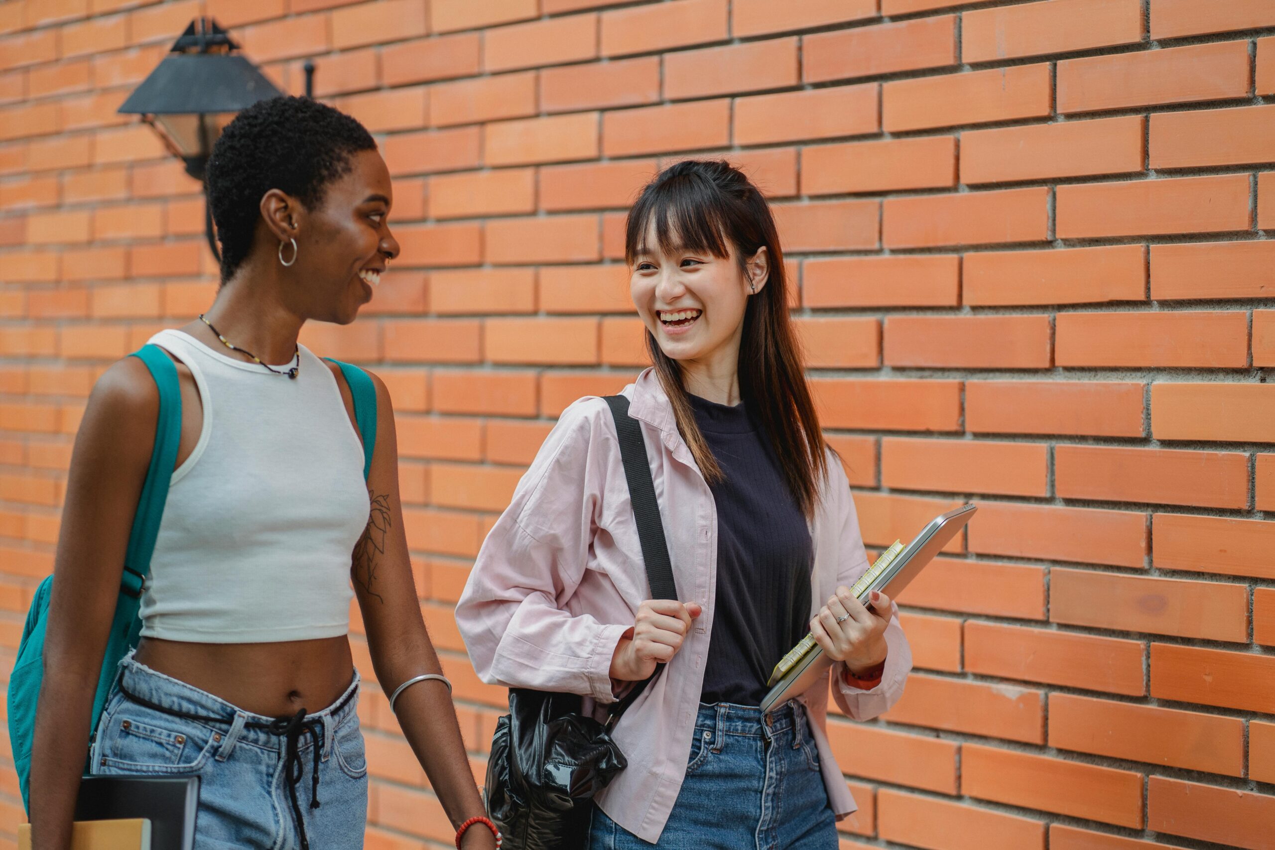Zwei junge Frauen gehen lachend vor einer Backsteinmauer. Eine hat kurze Haare, trägt ein bauchfreies Top und Jeans und trägt einen Rucksack. Die andere hat lange Haare, trägt eine leichte Jacke und hält einen Laptop und einige Bücher in der Hand. Beide scheinen in ein fröhliches Gespräch vertieft zu sein.