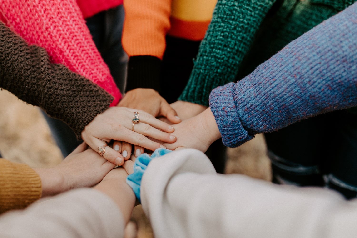 Eine Gruppe von Menschen in bunten Pullovern steht in einem Kreis und legt ihre Hände in der Mitte zusammen, was Einheit und Teamwork symbolisiert. Der Fokus liegt auf den Händen und der Vielfalt der Pullovertexturen.