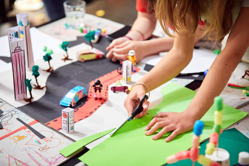 Kinder gestalten mit Schere und buntem Papier eine Miniatur-Stadtlandschaft mit Gebäuden, Bäumen, Autos und Figuren aus Papier, die auf einem Tisch angeordnet sind. Ihre Hände sind beim Basteln sichtbar.