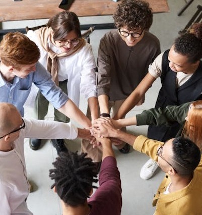 Eine Gruppe unterschiedlicher Menschen steht lächelnd in einem Kreis und legt in der Mitte ihre Hände zusammen, um in einer entspannten Innenumgebung Teamwork und Zusammenhalt zu demonstrieren.