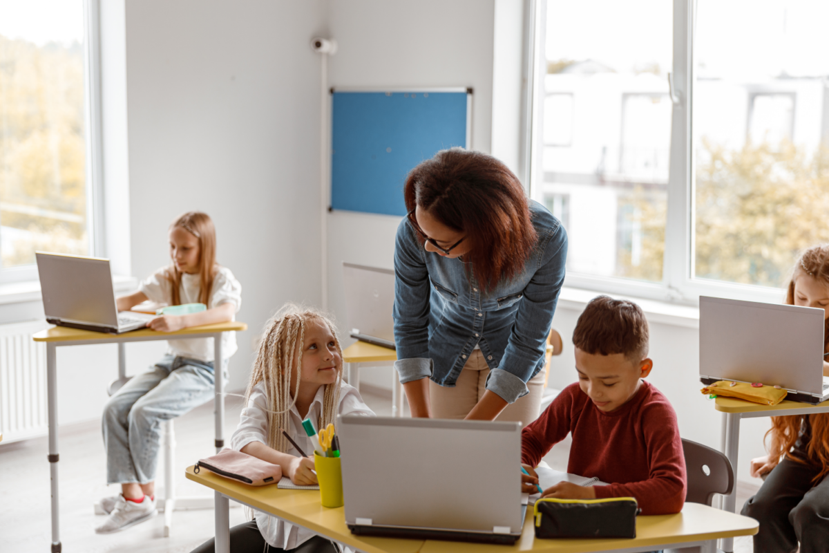 Ein Lehrer im Jeanshemd hilft einem jungen Schüler mit einem Laptop an einem Klassentisch. Andere Schüler, die ebenfalls Laptops benutzen, sitzen an gelben Tischen in einem hellen Raum mit großen Fenstern.