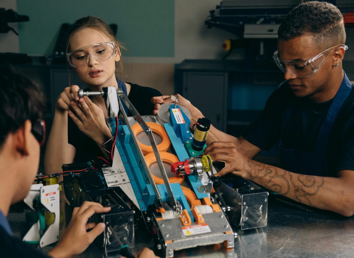 Drei Personen mit Schutzbrillen arbeiten gemeinsam am Bau eines Roboters. Sie konzentrieren sich auf das Zusammensetzen von Teilen und den Einsatz von Werkzeugen an einer Werkbank, was auf Teamwork und technische Fähigkeiten schließen lässt.
