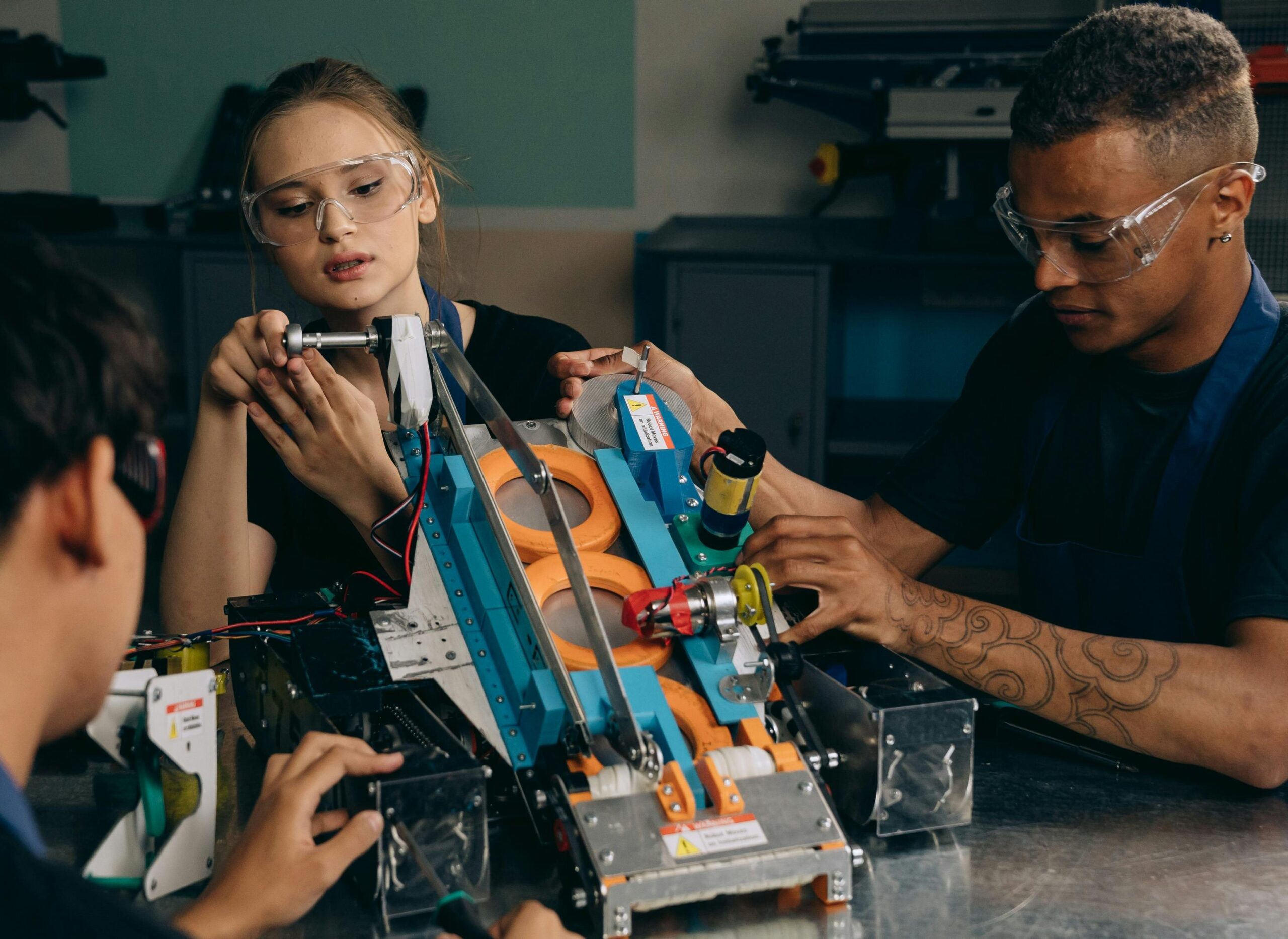 Drei Personen mit Schutzbrillen arbeiten gemeinsam am Bau eines Roboters. Sie konzentrieren sich auf das Zusammensetzen von Teilen und den Einsatz von Werkzeugen an einer Werkbank, was auf Teamwork und technische Fähigkeiten schließen lässt.