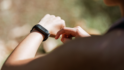 Eine Person tippt im Freien auf eine Smartwatch. Der verschwommene Hintergrund aus Gras und Blättern suggeriert eine Natur- oder Parkumgebung. Das Armband ist schwarz und die Haut der Person hat einen hellen Hautton.