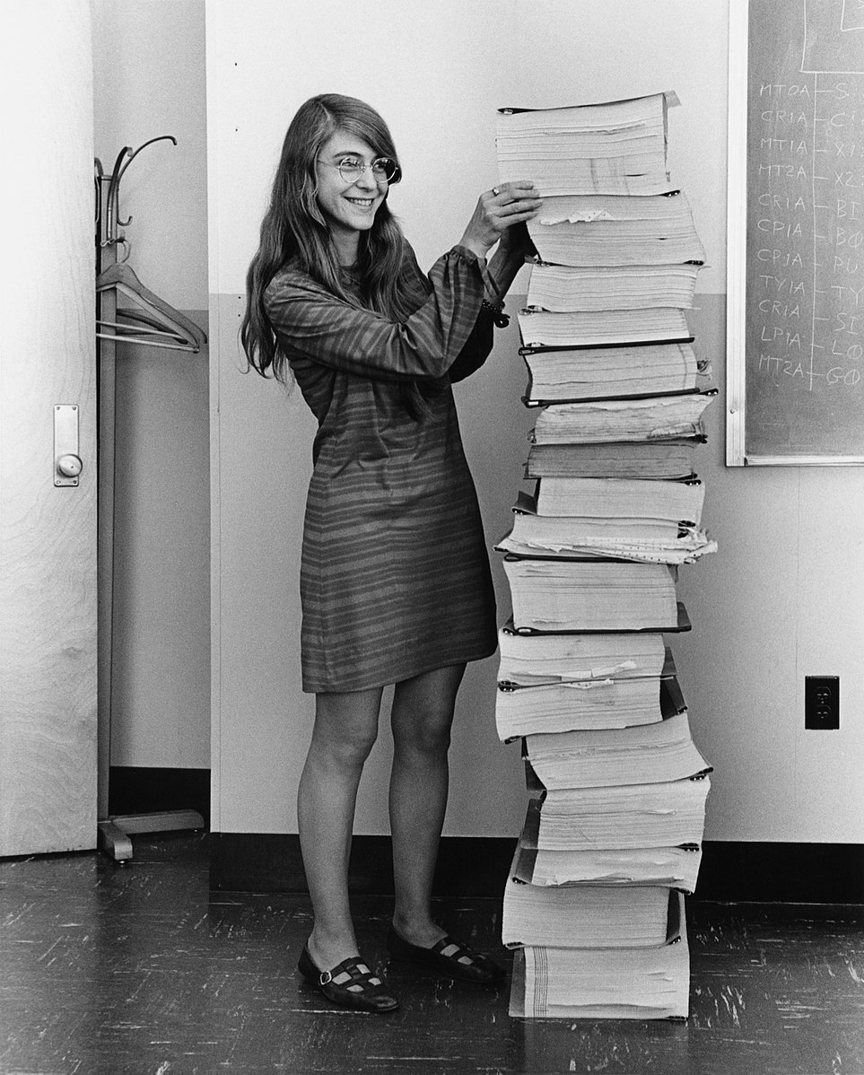 Margaret Hamilton in gestreiftem Kleid und mit Brille steht lächelnd neben einem hohen Stapel großer, dicker Computerausdrucke. Sie hält den Stapel fest, damit er nicht umfällt. Im Hintergrund sind eine Tafel und eine Garderobe zu sehen.