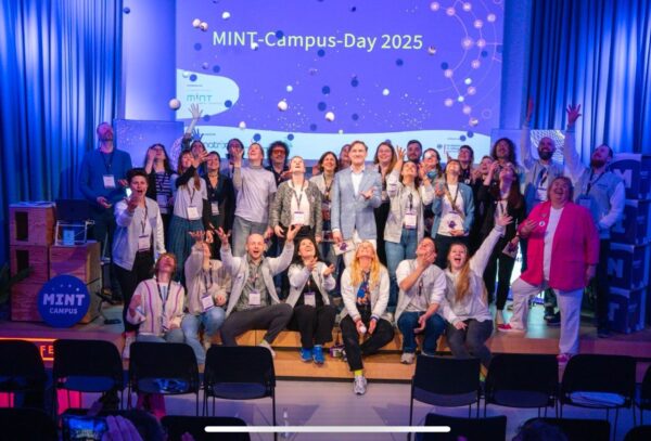Eine Gruppe von Menschen aus der MINT-Community posiert fröhlich auf der Bühne des „MINT-Campus-Tages 2025“. Einige werfen ihre Hände in die Luft und stehen vor einem Bildschirm mit dem Veranstaltungsnamen. Überall auf der Bühne sind Requisiten verteilt.