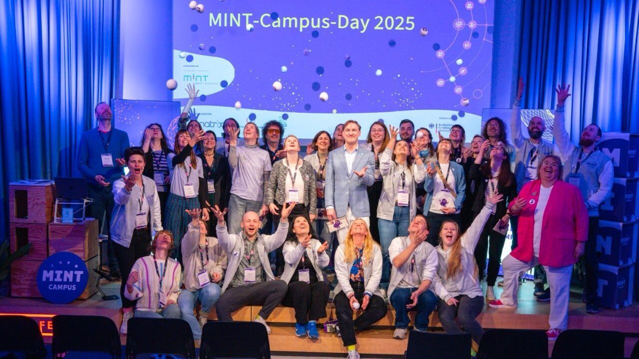 Eine Gruppe von Menschen aus der MINT-Community posiert fröhlich auf der Bühne des „MINT-Campus-Tages 2025“. Einige werfen ihre Hände in die Luft und stehen vor einem Bildschirm mit dem Veranstaltungsnamen. Überall auf der Bühne sind Requisiten verteilt.