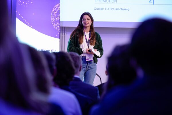 Lisa Graf steht auf der Bühne des MINT-Campus und spricht zum Publikum. Sie trägt eine grüne Jacke und Jeans vor einem violett gestalteten Hintergrund mit teilweise sichtbarem Text auf der Leinwand. Das Publikum ist als Silhouette von hinten zu sehen.