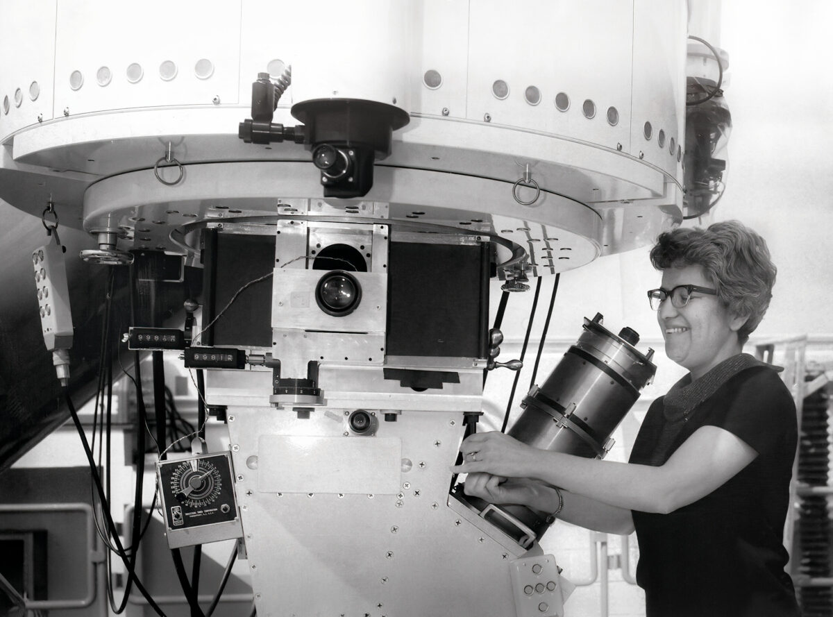 Eine Frau mit Brille lächelt, während sie in einem Labor mit einem großen, komplexen wissenschaftlichen Instrument arbeitet und möglicherweise mithilfe moderner Observatoriumsausrüstung Dunkle Materie erforscht.