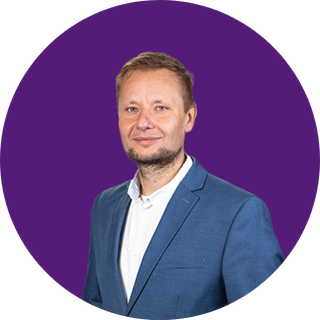 Ein Mann mit kurzen hellbraunen Haaren und einem gestutzten Bart trägt eine blaue Anzugsjacke und ein weißes Hemd und steht vor einem lilafarbenen, kreisförmigen Hintergrund, der das professionelle Team hinter unserer Über uns-Seite darstellt.